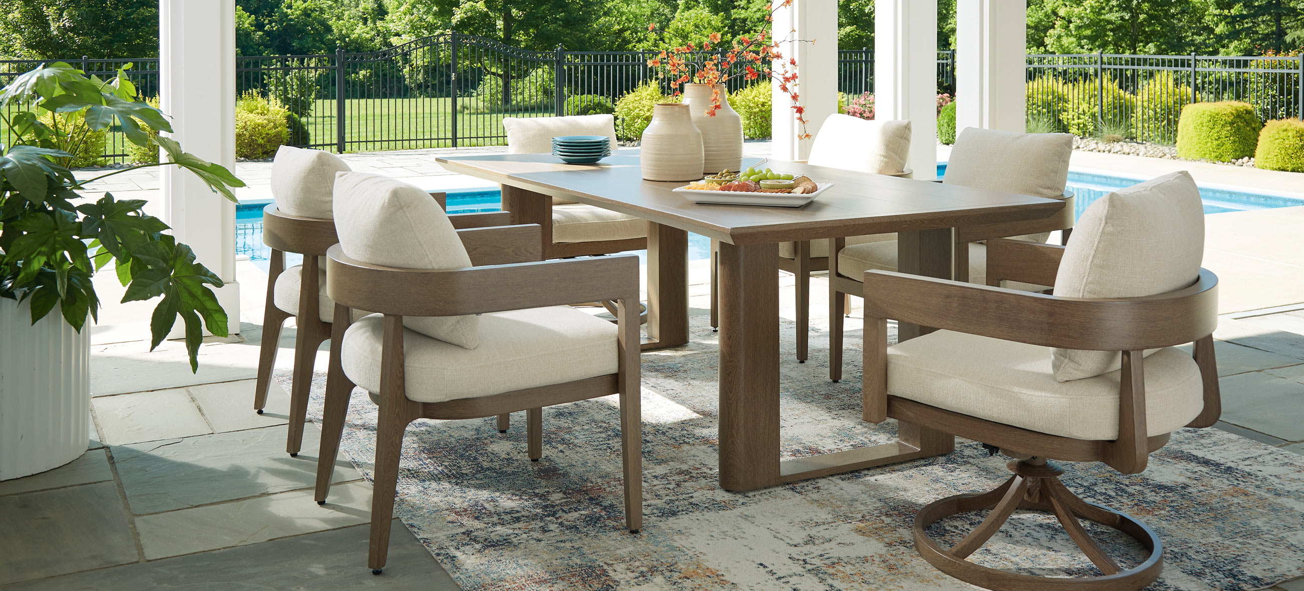 A stylish outdoor Serene Bay dining set features a wooden table and cushioned chairs, surrounded by greenery and a pool in a sunny setting.