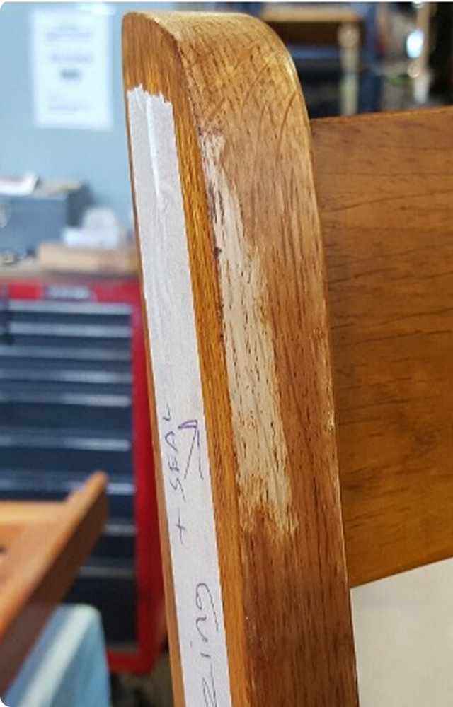 Close-up of a wooden chair's armrest with wear and scratches. A paper taped on it has handwritten notes. A “Before” label is in the top left corner.