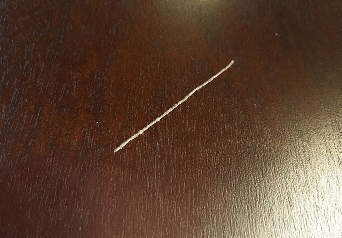 A dark wooden surface with a noticeable, white scratch running diagonally across the center. The tone is blemished and slightly worn.