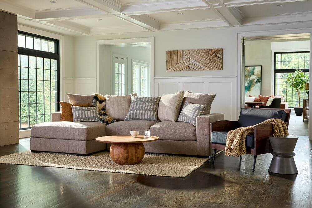 A cozy living room with a beige sectional sofa, patterned cushions, a soft ottoman, and a multicolored rug. Natural light pours through large windows.