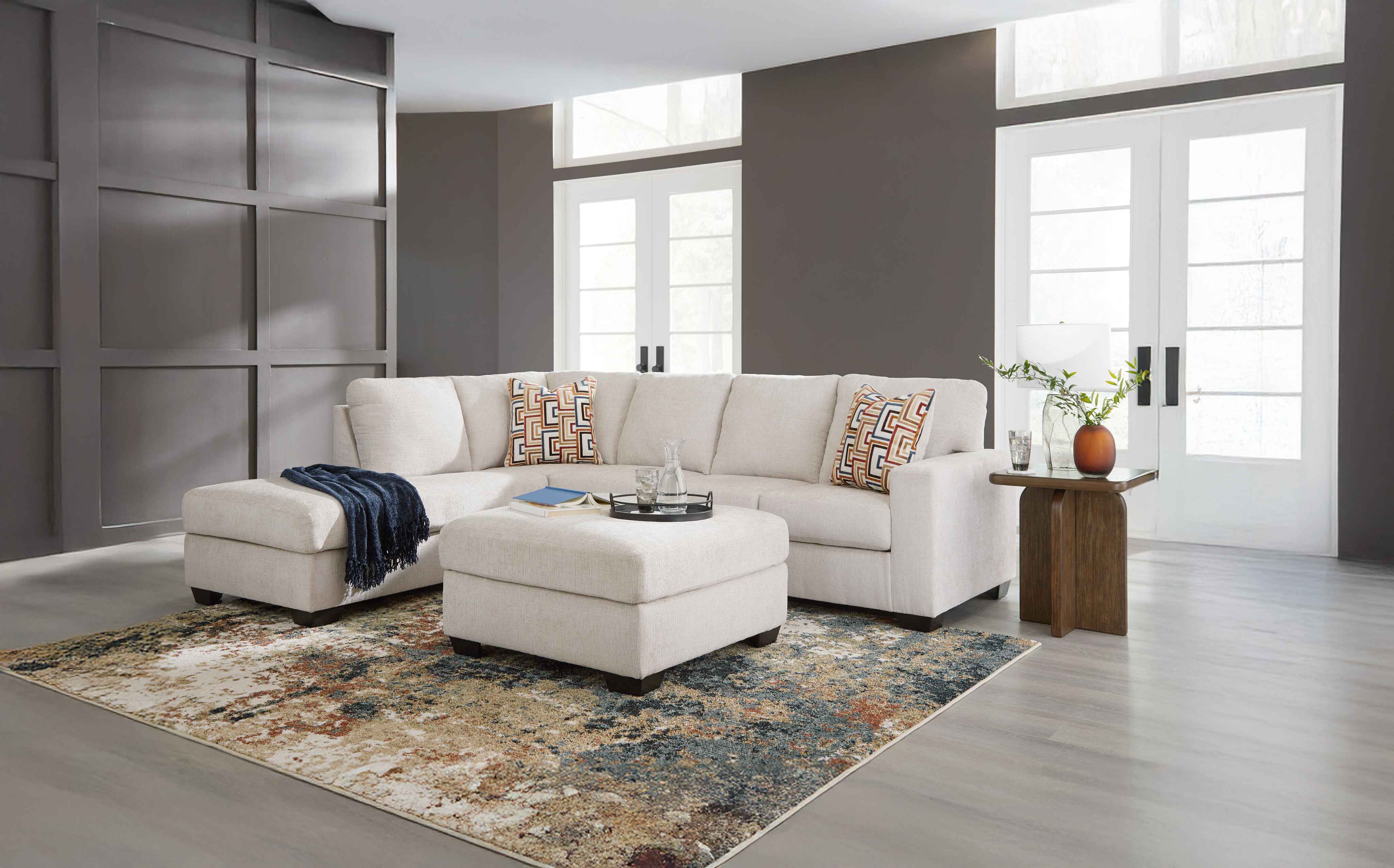 A cozy living room with a beige sectional sofa, patterned cushions, a soft ottoman, and a multicolored rug. Natural light pours through large windows.