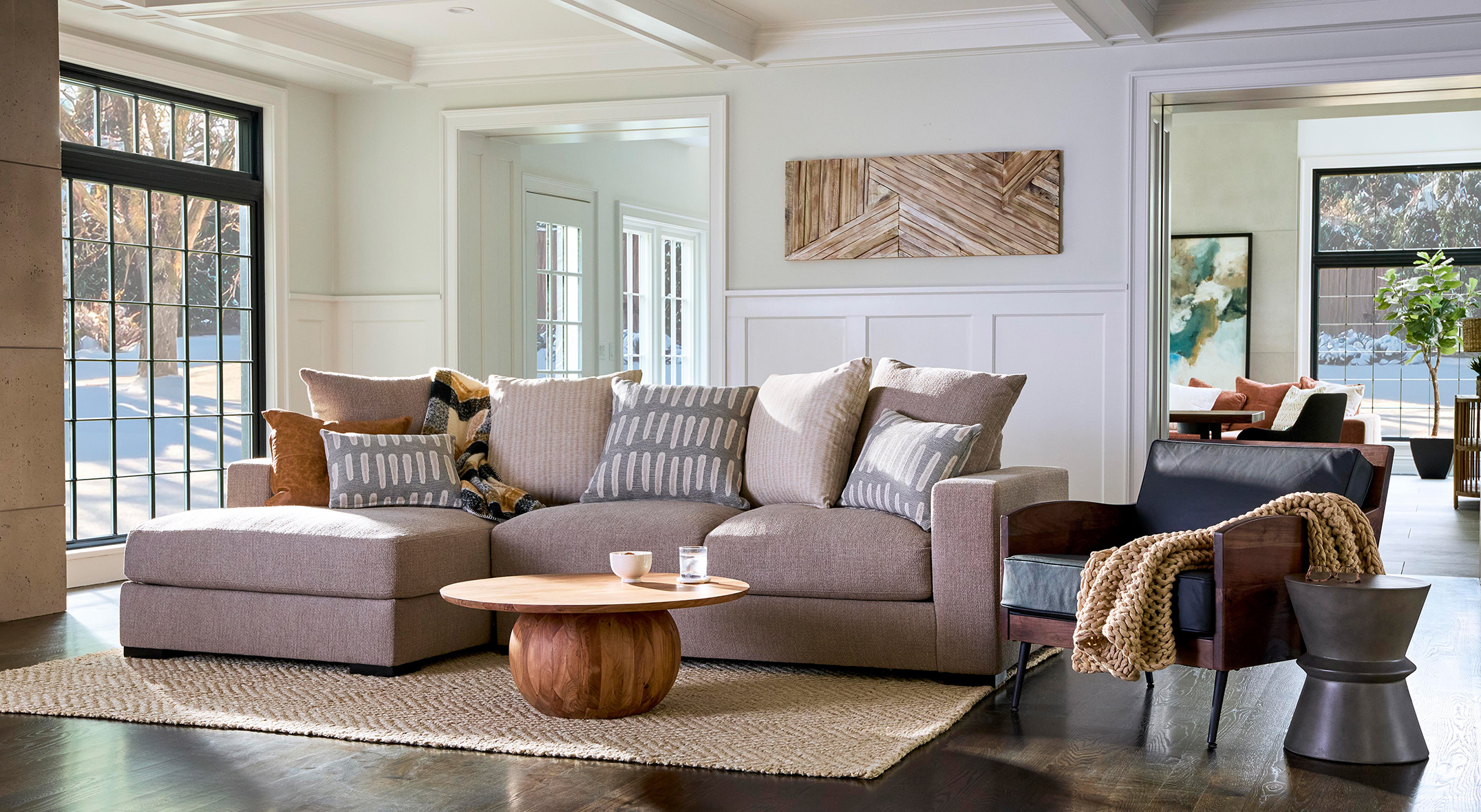 A cozy living room with a beige sectional sofa, patterned cushions, a soft ottoman, and a multicolored rug. Natural light pours through large windows.