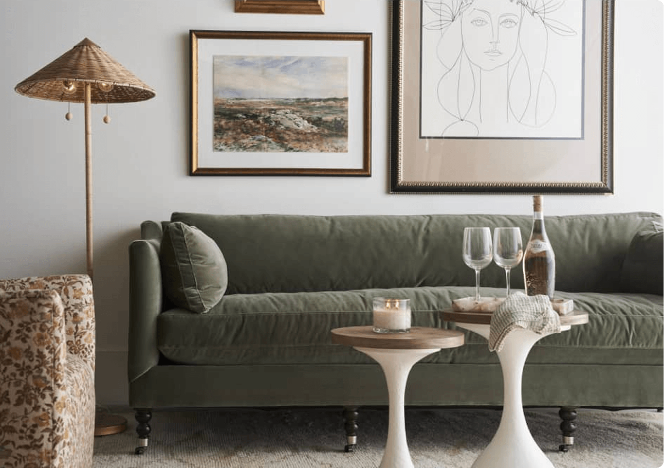 A serene living room with an olive-green sofa, framed landscape and line-art portraits, woven floor lamp, and petite tables set with wine glasses.