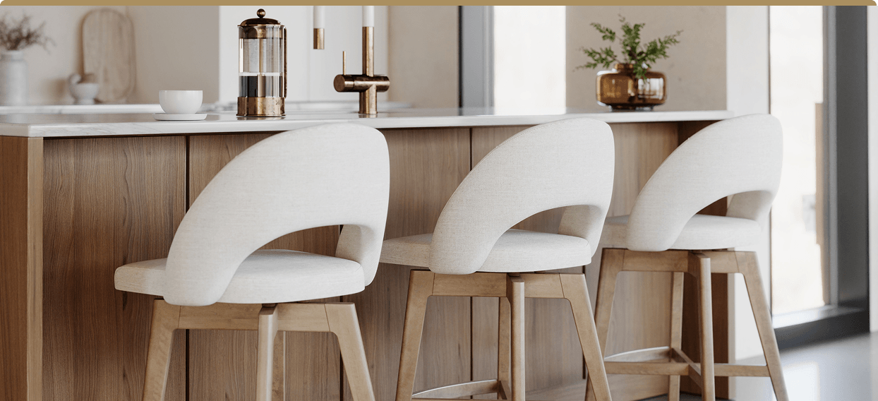 Three white upholstered bar stools with wooden legs 
            line a warm wood kitchen island, beside a French press and small potted plant in soft natural light.