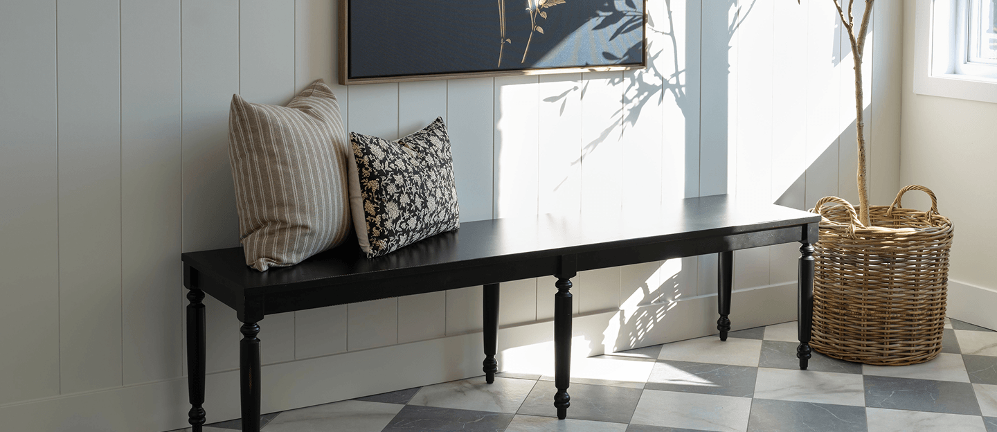 A black console bench against pale paneled walls holds two textured cushions—a neutral striped pillow and a floral one—creating a calm, elegant interior.