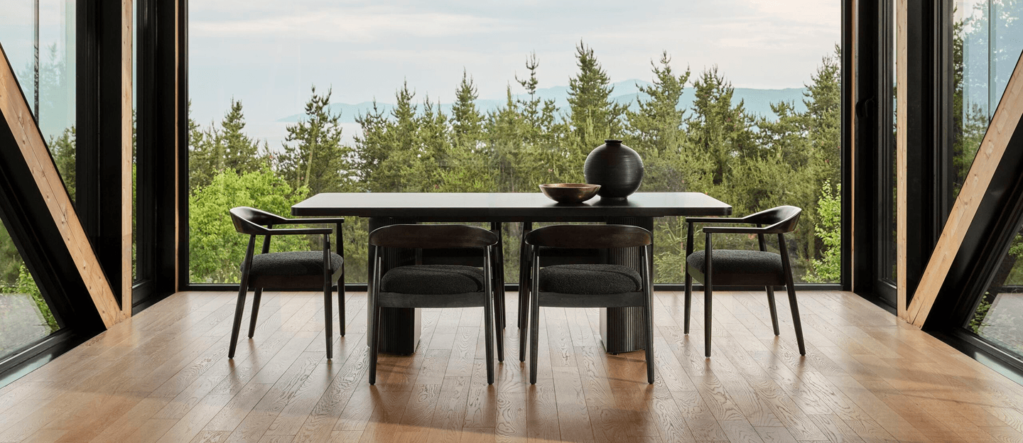 A minimalist wooden dining table and chairs sit in a glass-walled room overlooking a lush forest, creating a calm, airy atmosphere.
