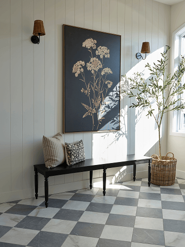 A black console bench against pale paneled walls holds two textured cushions—a neutral striped pillow and a floral one—creating a calm, elegant interior.