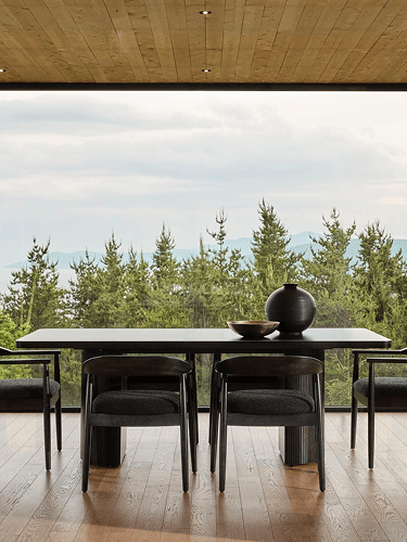 A minimalist wooden dining table and chairs sit in a glass-walled room overlooking a lush forest, creating a calm, airy atmosphere.