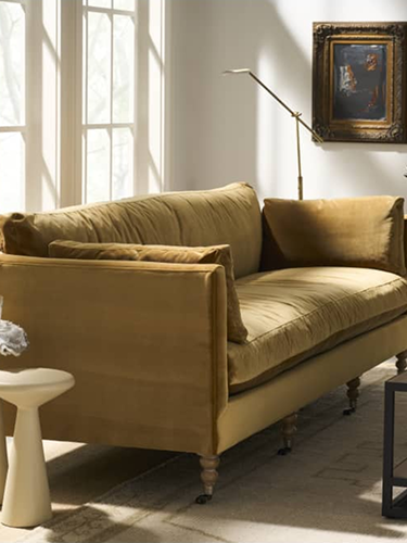 Sunlit living room with a tan sofa, white side table, leafy plant, and dark coffee table; tall windows create a calm, inviting atmosphere.