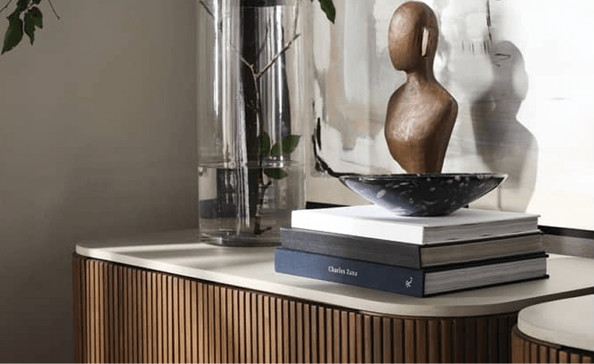 A wooden bust, black bowl, art books and leafy branches style a ribbed console beneath abstract art, creating a calm, modern interior.