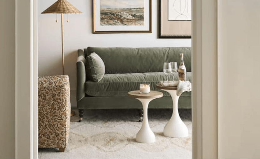 A serene living room features a deep green sofa, sculptural white tables, a patterned ottoman, a floor lamp, and framed art in warm neutrals.