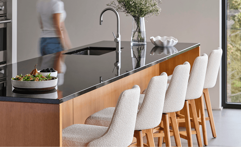 Bright modern kitchen with a long wood-and-stone island, four upholstered stools, a curved faucet, flowers, and a blurred person walking nearby; calm, airy feel.