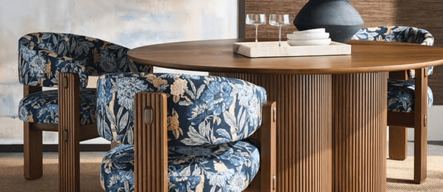 A warm modern dining room features a round wood table, patterned blue chairs, a sculptural chandelier, abstract art, and greenery.
