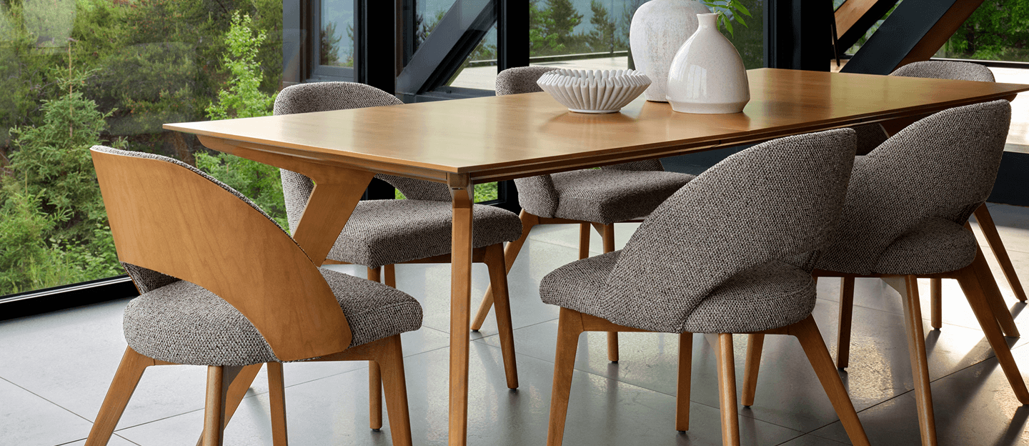 A warm modern dining area with a light wood table, sculptural gray chairs, ceramic decor, and large windows overlooking lush greenery.