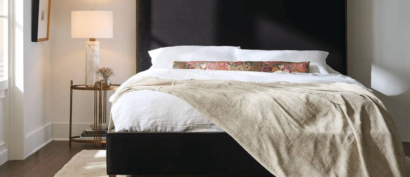 A neatly made bed with white linens and a soft beige throw sits against a dark headboard, beside a lamp and flowers for a calm, modern feel.