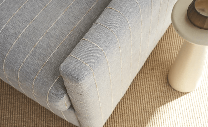 Close-up of a light gray upholstered sofa with rounded, 
                    stitched seams on a woven beige rug, beside a cream-colored lamp; the scene feels calm and refined.