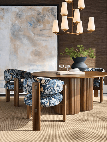 A warm modern dining room features a round wood table, patterned blue chairs, a sculptural chandelier, abstract art, and greenery.