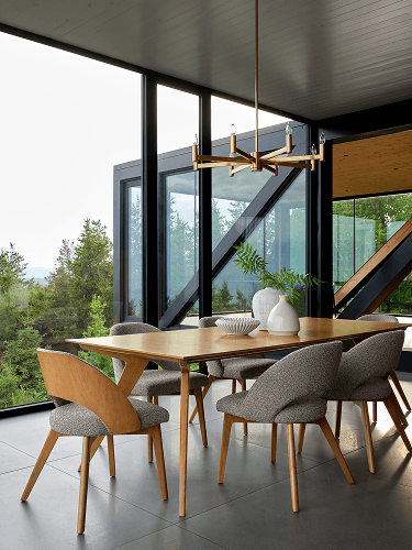 A warm modern dining area with a light wood table, sculptural gray chairs, ceramic decor, and large windows overlooking lush greenery.