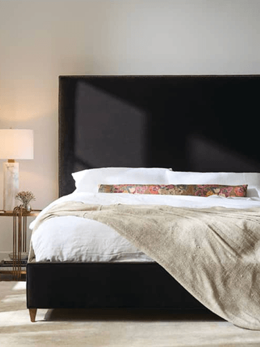 A neatly made bed with white linens and a soft beige throw sits against a dark headboard, beside a lamp and flowers for a calm, modern feel.