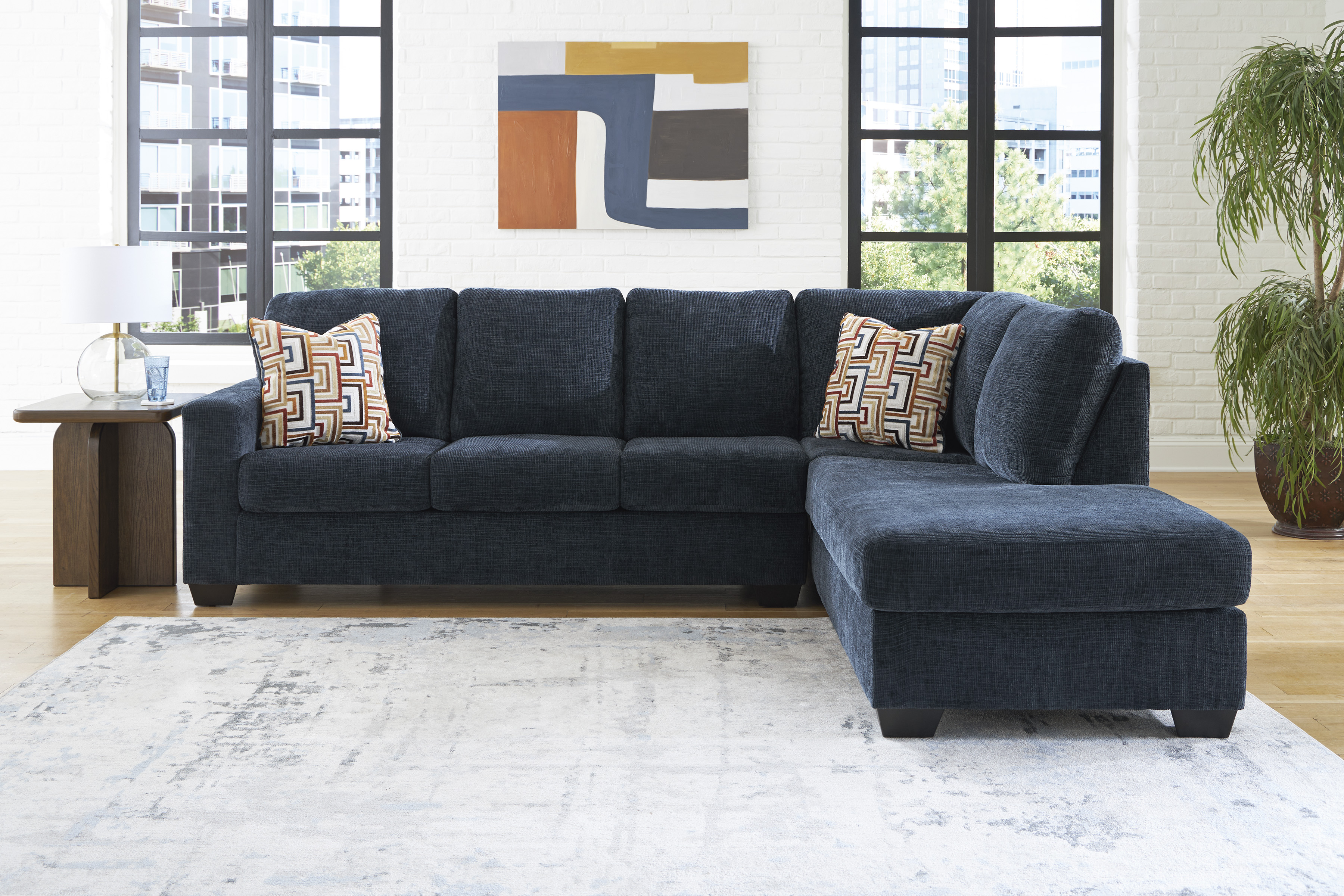 modern living room with a cream sectional sofa, patterned pillows, ottoman, blue throw, abstract art, plants, and a textured rug