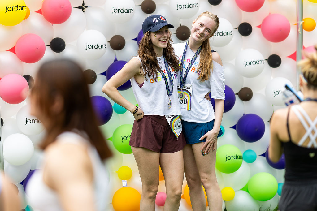 Two smiling women in athletic gear pose with medals in front of a colorful balloon wall. They exude joy, celebrating an event with a casual, festive vibe.
