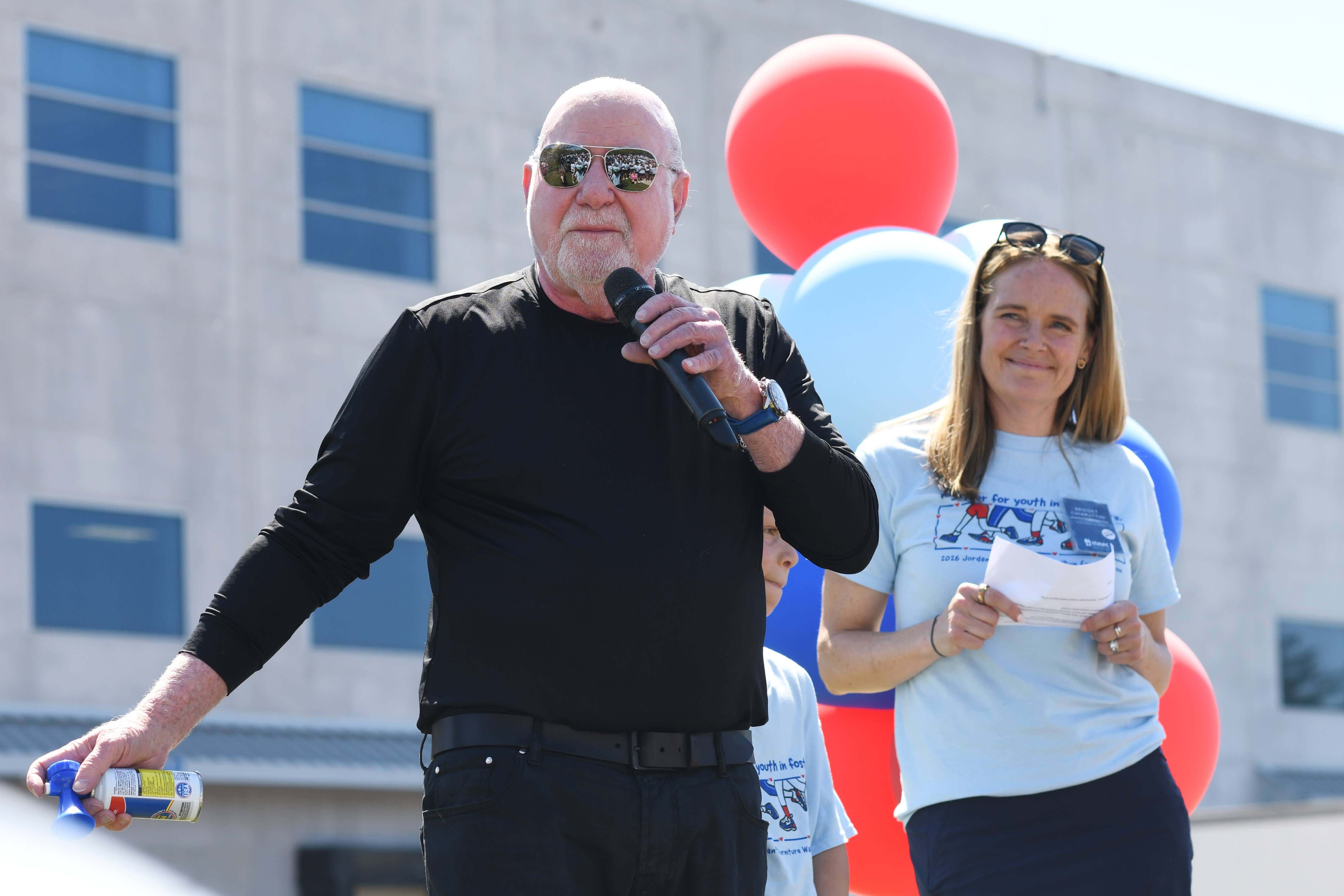 Eliot Tatelman speaks into a microphone, smiling next to a woman holding papers. They stand in front of red and blue balloons outside a building.