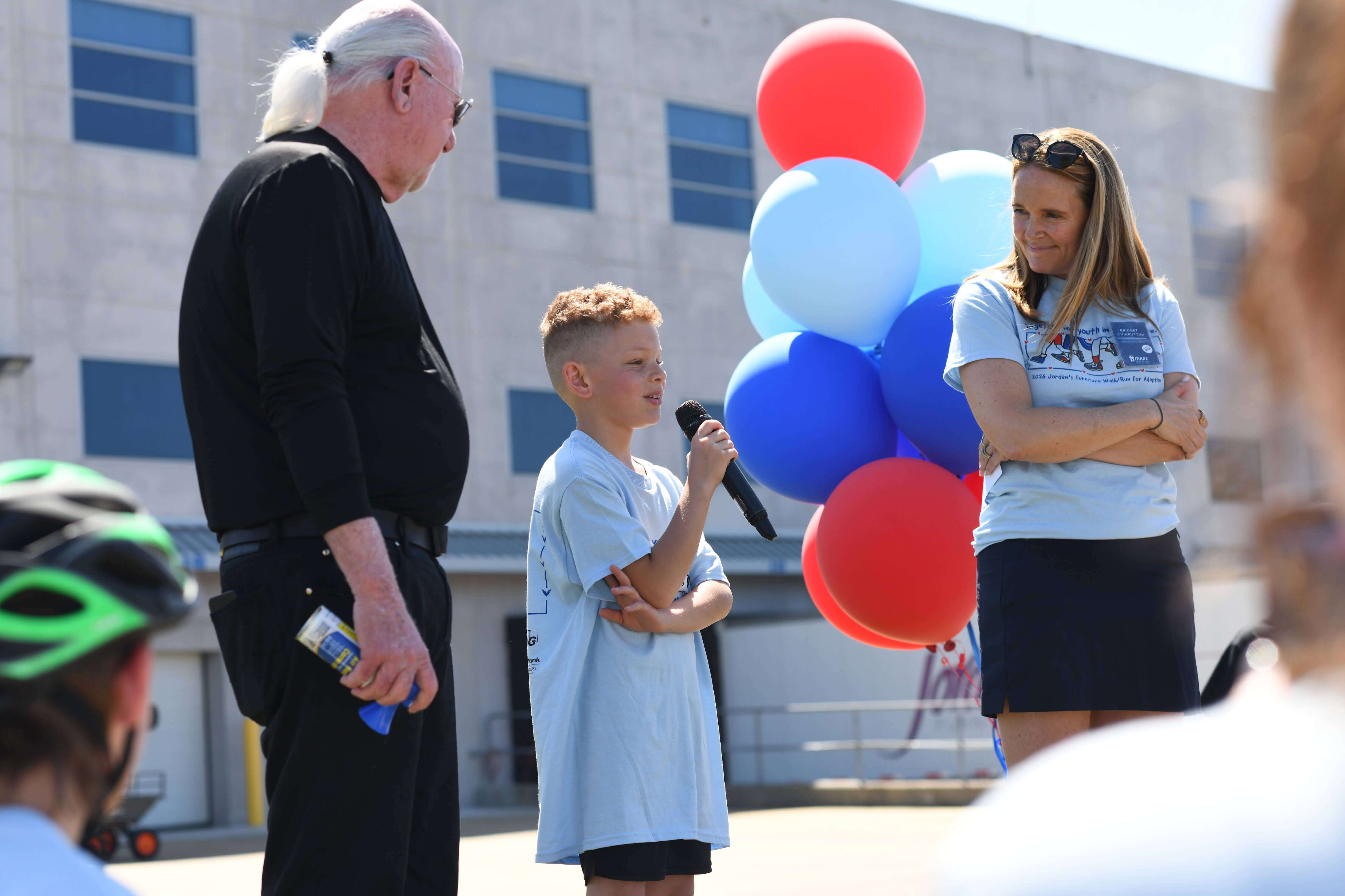 A young boy speaks into a microphone outdoors, flanked by Eliot Tatelman and a woman. Behind them are red and blue balloons. The atmosphere is lively and sunny.