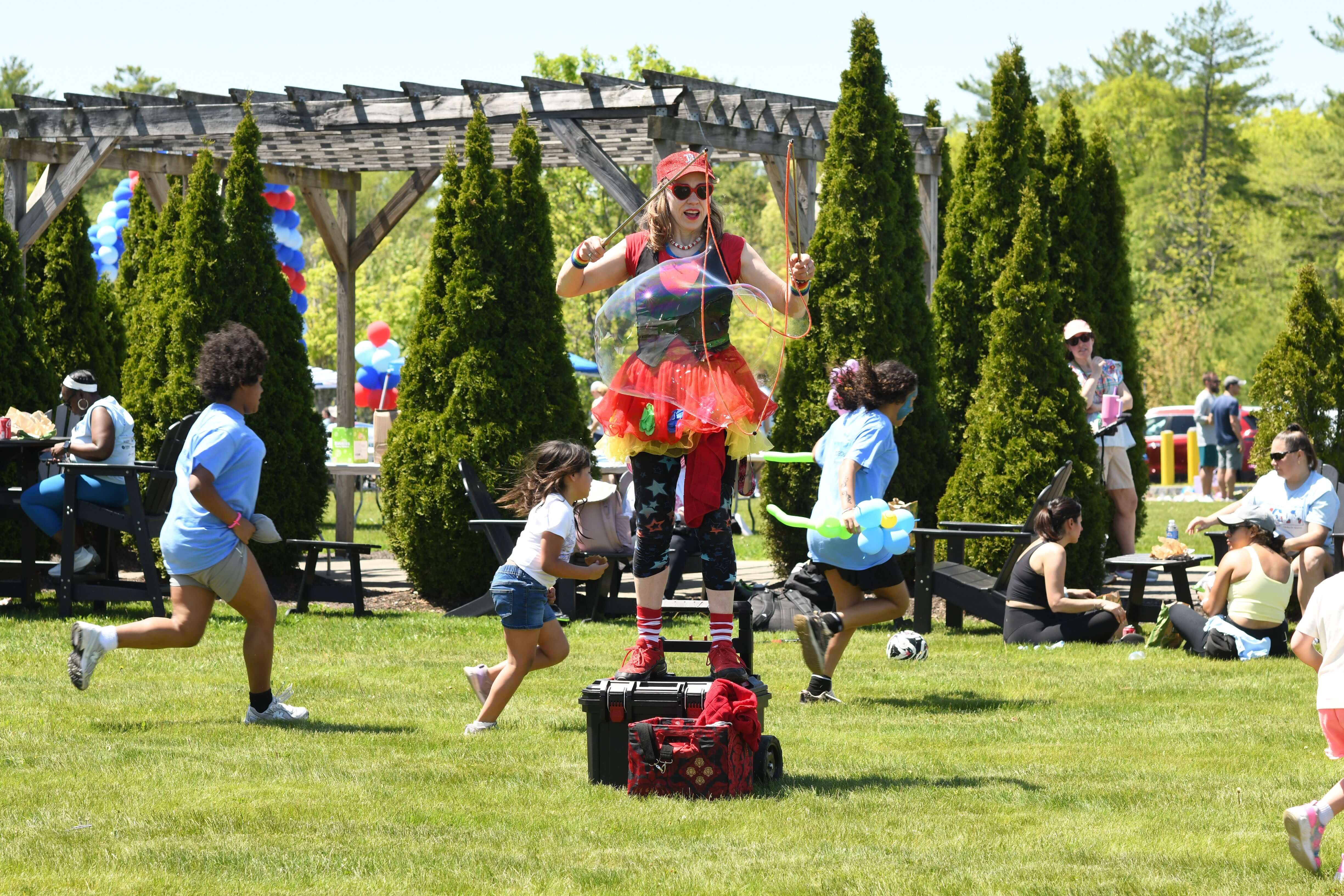 A performer on stilts wearing a colorful outfit entertains children with bubbles in a sunny park, evoking joy and excitement.