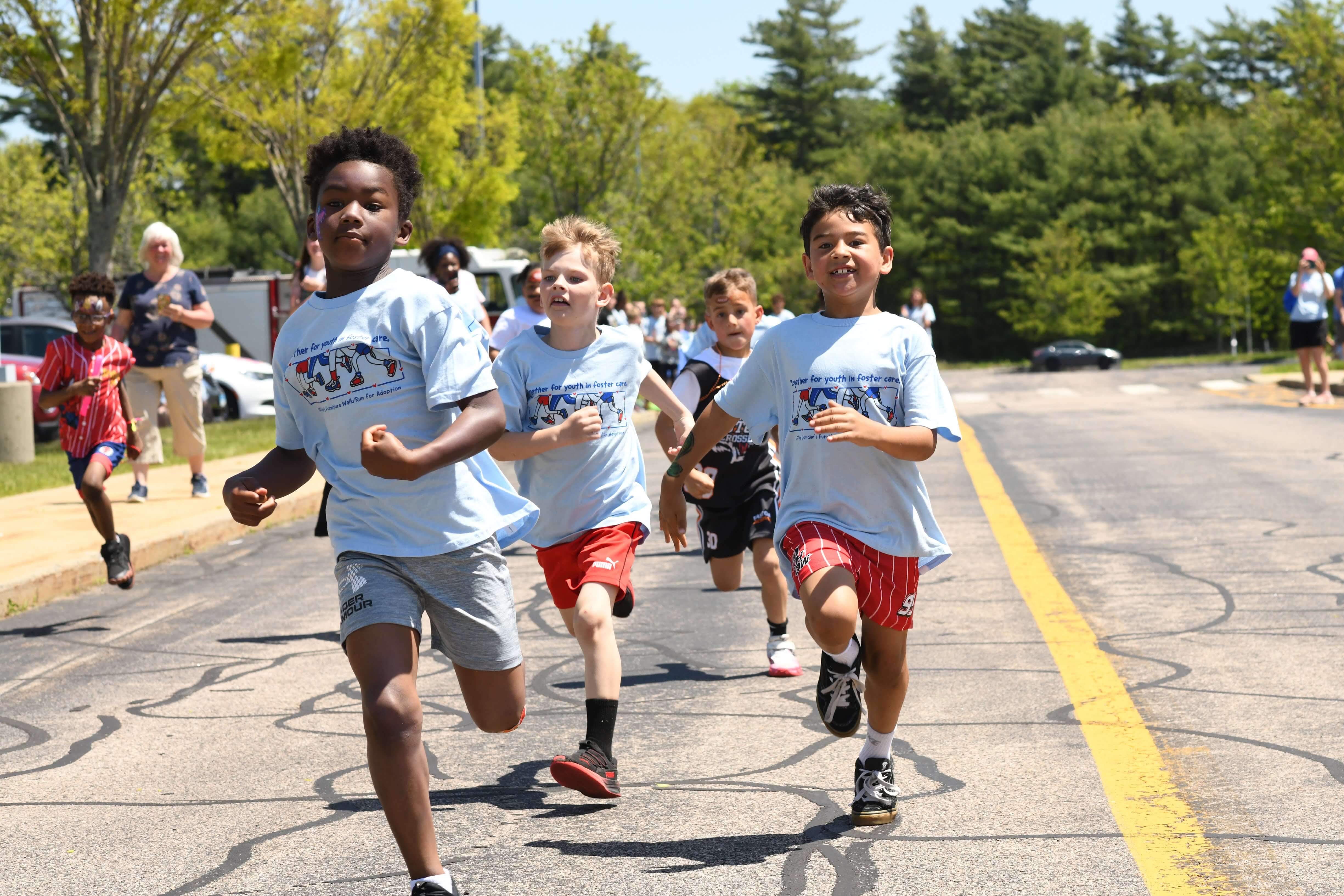 Children participate in a joyful outdoor race on a sunny day, wearing matching light blue shirts. Trees and spectators line the background.