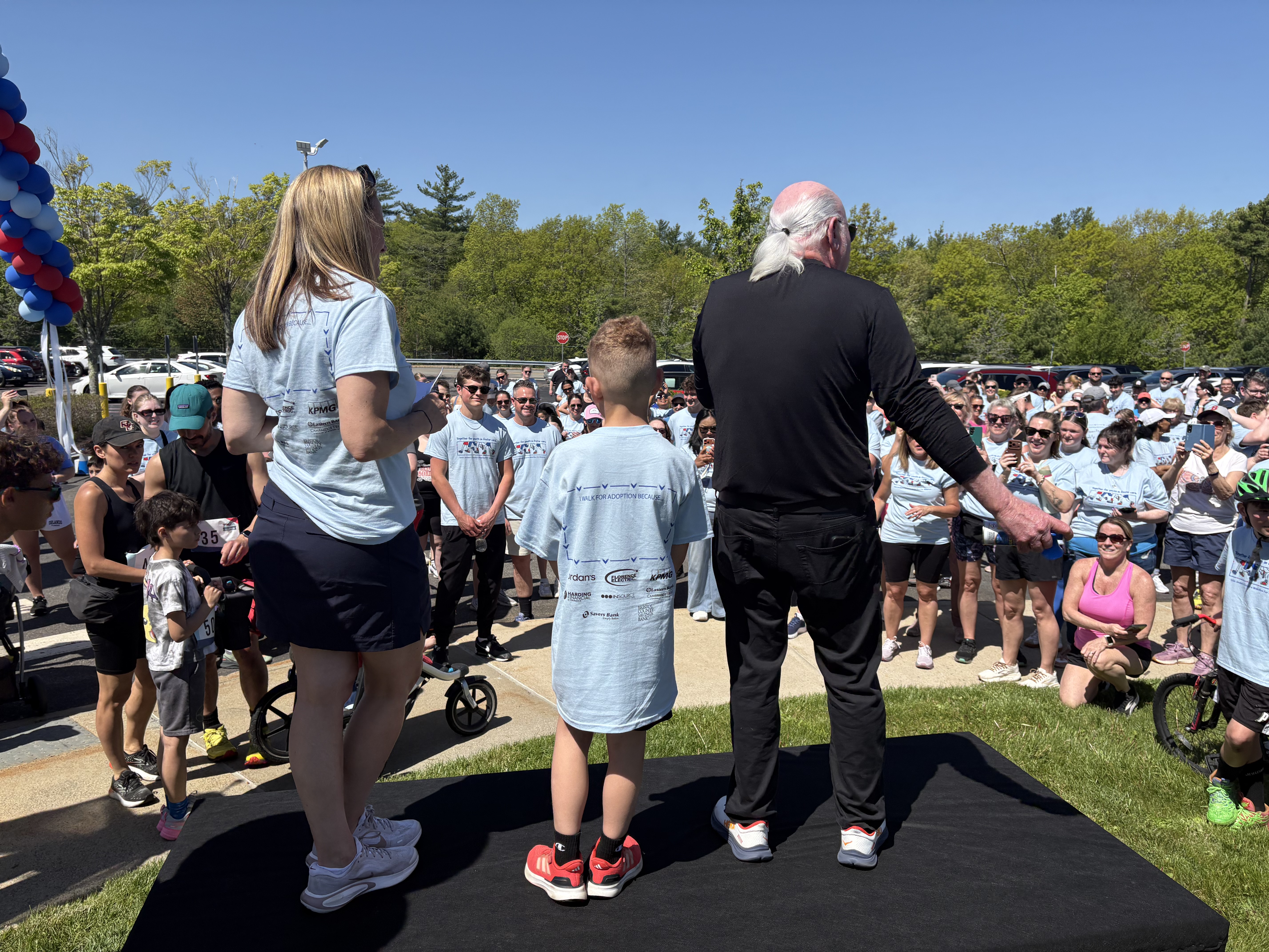 Eliot Tatelman and boy stand onstage facing a crowd at an outdoor event under a clear sky. The audience, wearing matching blue shirts, gathers attentively.
