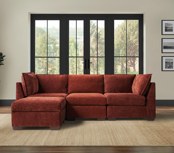 A cozy, rust-colored sectional sofa with two corners, one armless section, and an ottoman, set against large windows with a peaceful outdoor view.