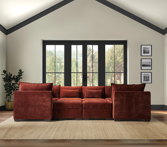 A cozy, rust-colored sectional sofa with two corners, three armless sections, and an ottoman, set against large windows with a peaceful outdoor view.