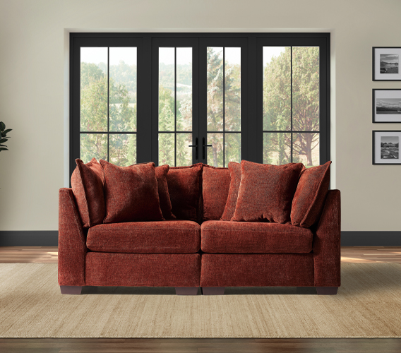 A 'two corner' cozy brown sofa with plush cushions is placed in front of large, black-framed windows revealing a lush green view. Black-and-white photos adorn the beige walls.