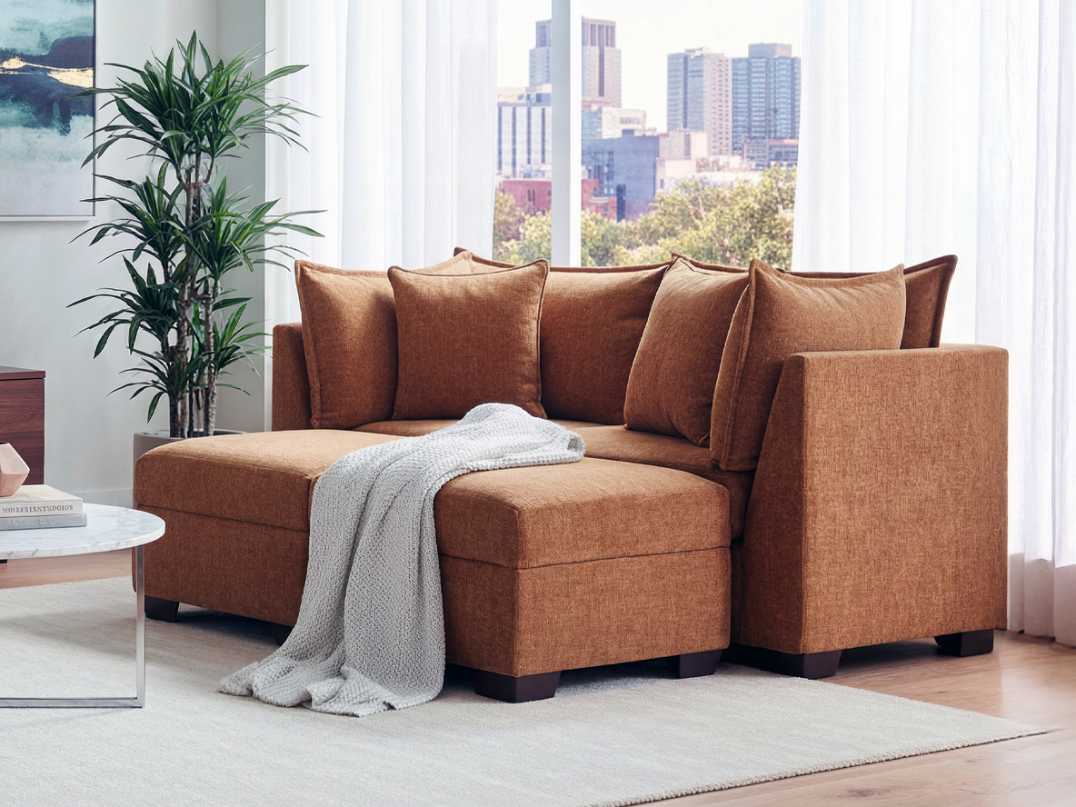 A cozy, mustard-colored sectional sofa with plush cushions is adorned with a gray knitted throw. It's set in a bright room with a potted plant and city view.