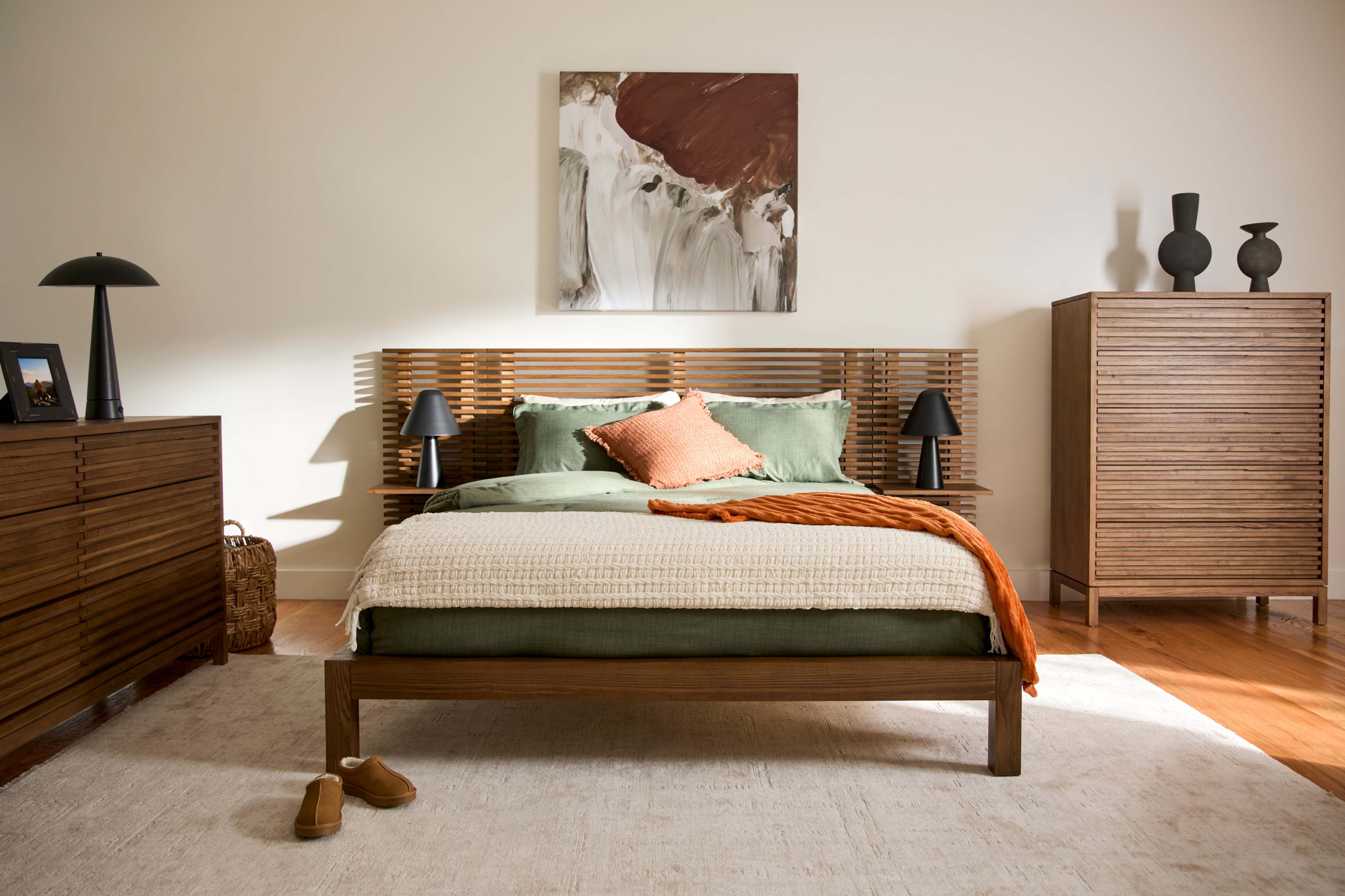 Warm mid-century modern bedroom with a neatly made bed, teal and rust pillows, wooden dressers, framed art, and sunlight across the floor.