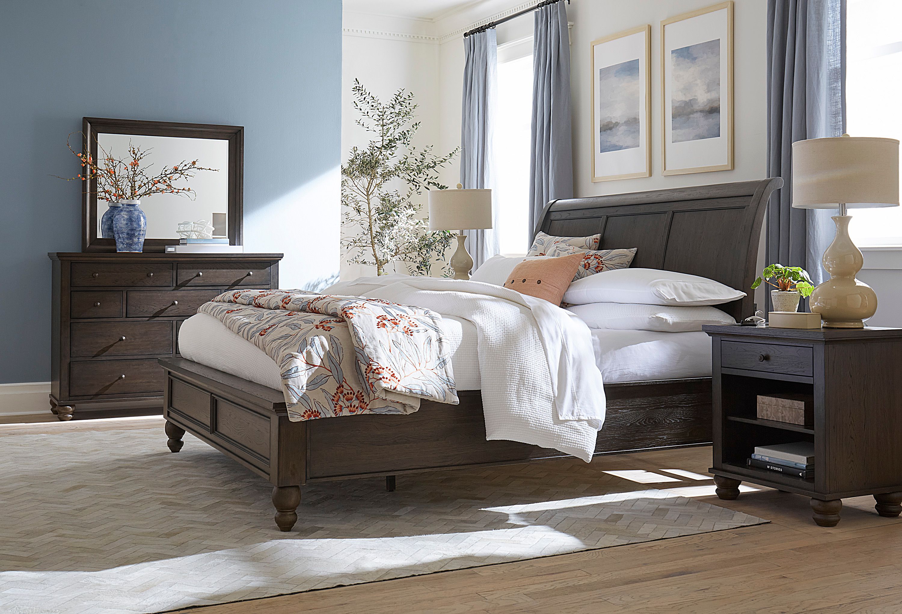 Sunlit modern bedroom with an upholstered bed, rumpled white-and-patterned bedding, gray bench, bedside tables, and soft neutral decor.