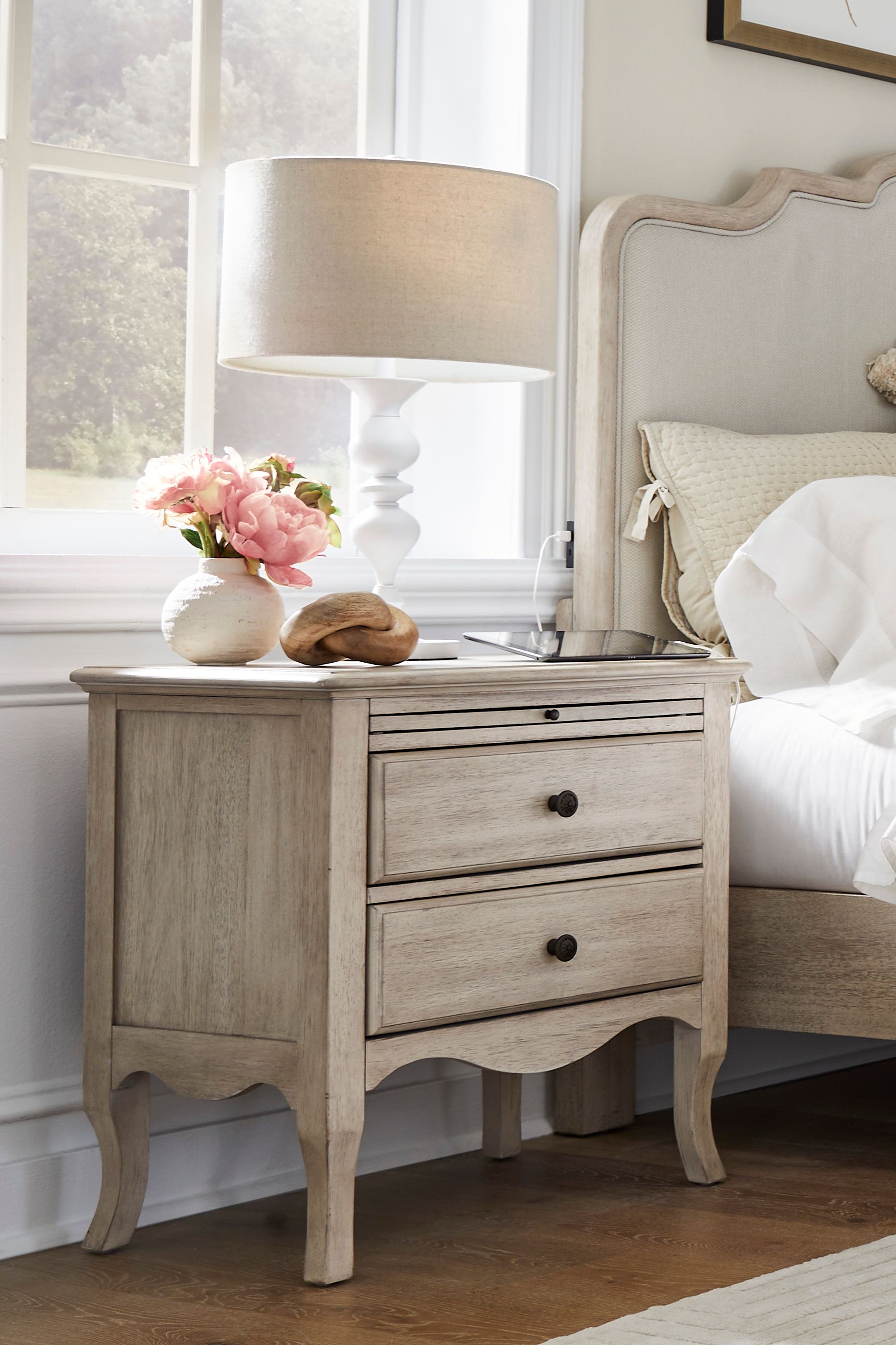 Bright, serene bedroom corner with a vintage white dresser, pink flowers, lamp, framed art, and upholstered bed in soft neutral tones.