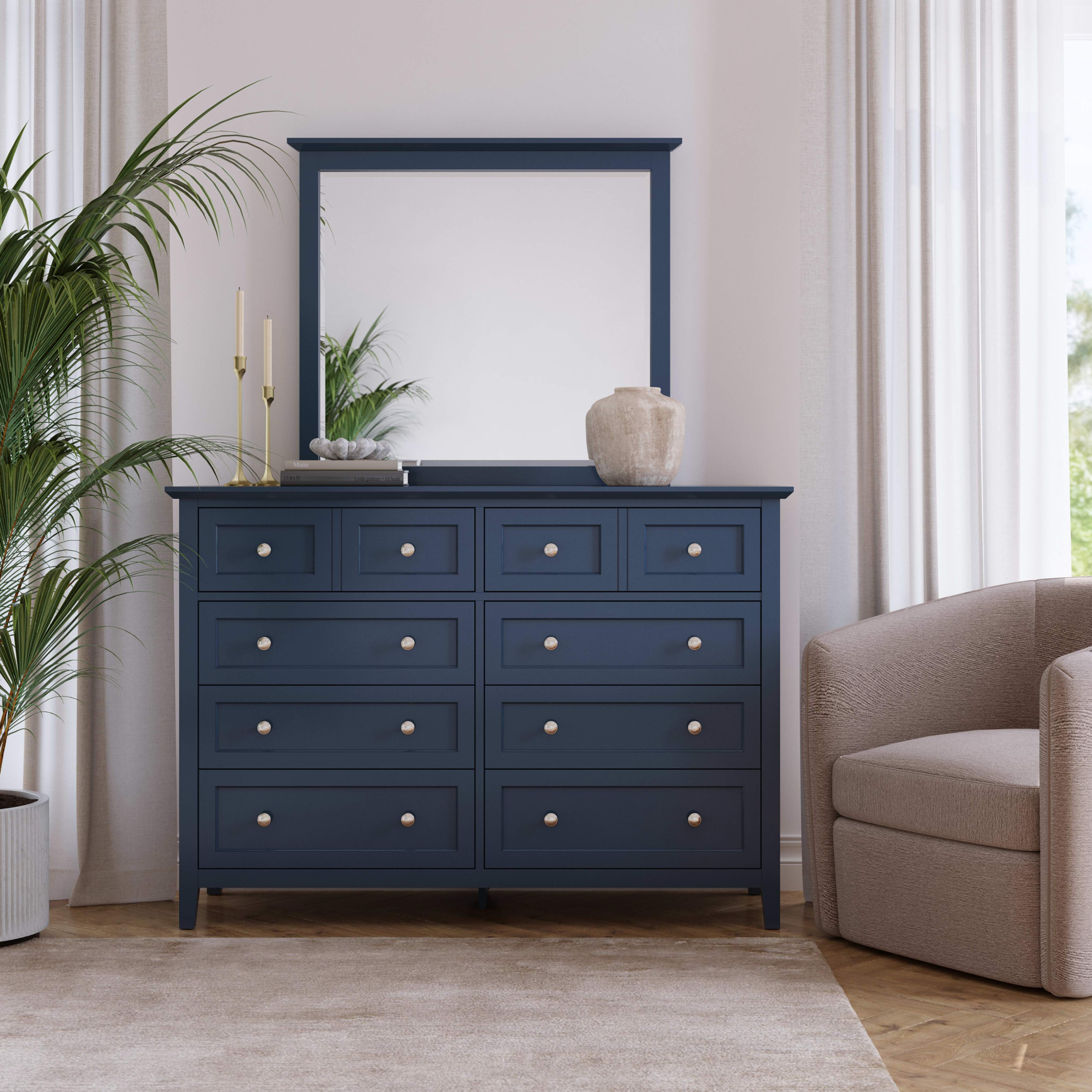 Bright, serene interior with a dark navy six-drawer dresser, leafy potted plants, beige armchair, and soft daylight through white curtains.