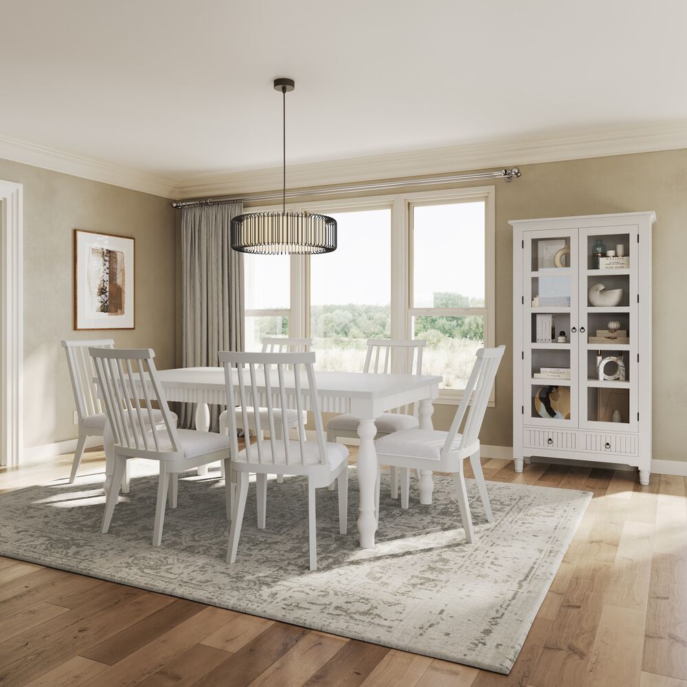 Sunlit dining room with a white table and six chairs on a patterned rug, a glass-front cabinet, large windows, and a modern pendant light. The neutral decor feels calm and airy.