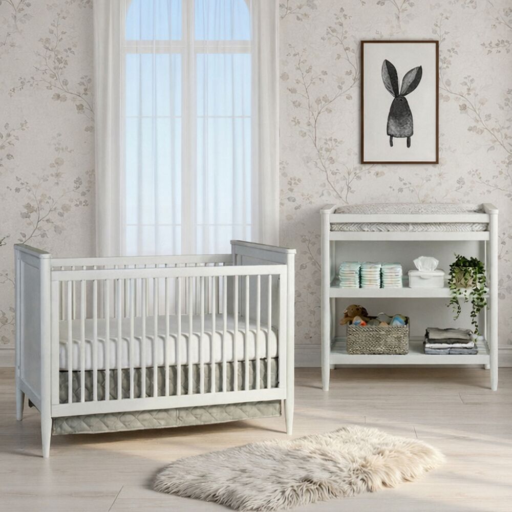 A calm, neutral nursery with a white crib beside a low dresser, framed monochrome print on the wall, pale patterned wallpaper, and a soft rug.