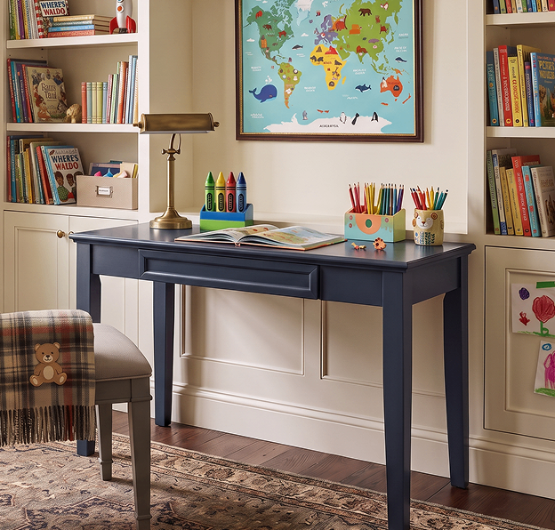 A cozy children’s study features a navy desk with an open book and colorful pencils, a world map, full bookshelves, and a teddy-bear chair.