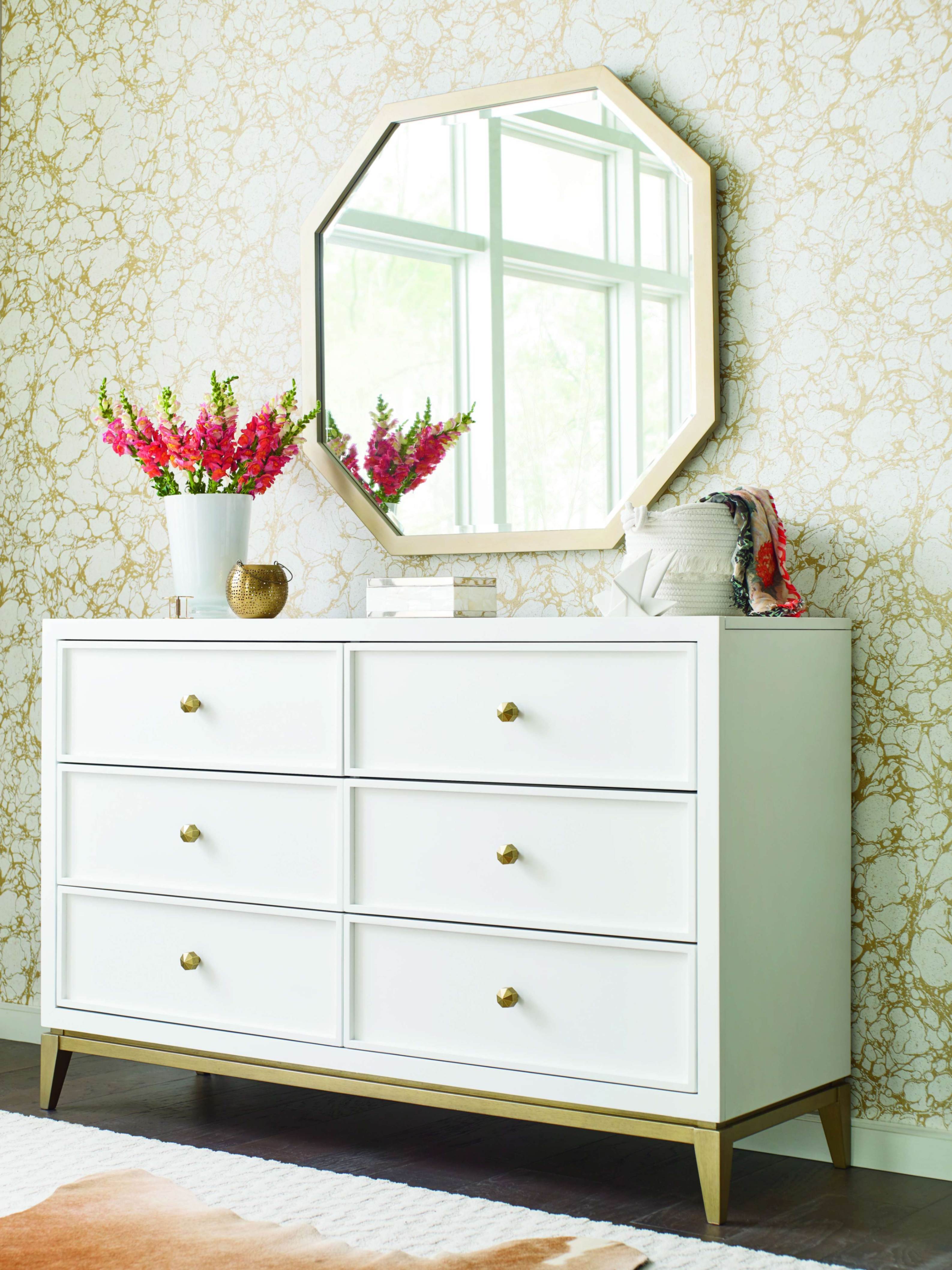 A bright bedroom vignette features a white six-drawer dresser with brass knobs beneath a geometric mirror, flowers, and soft patterned wallpaper, creating a calm, elegant mood.