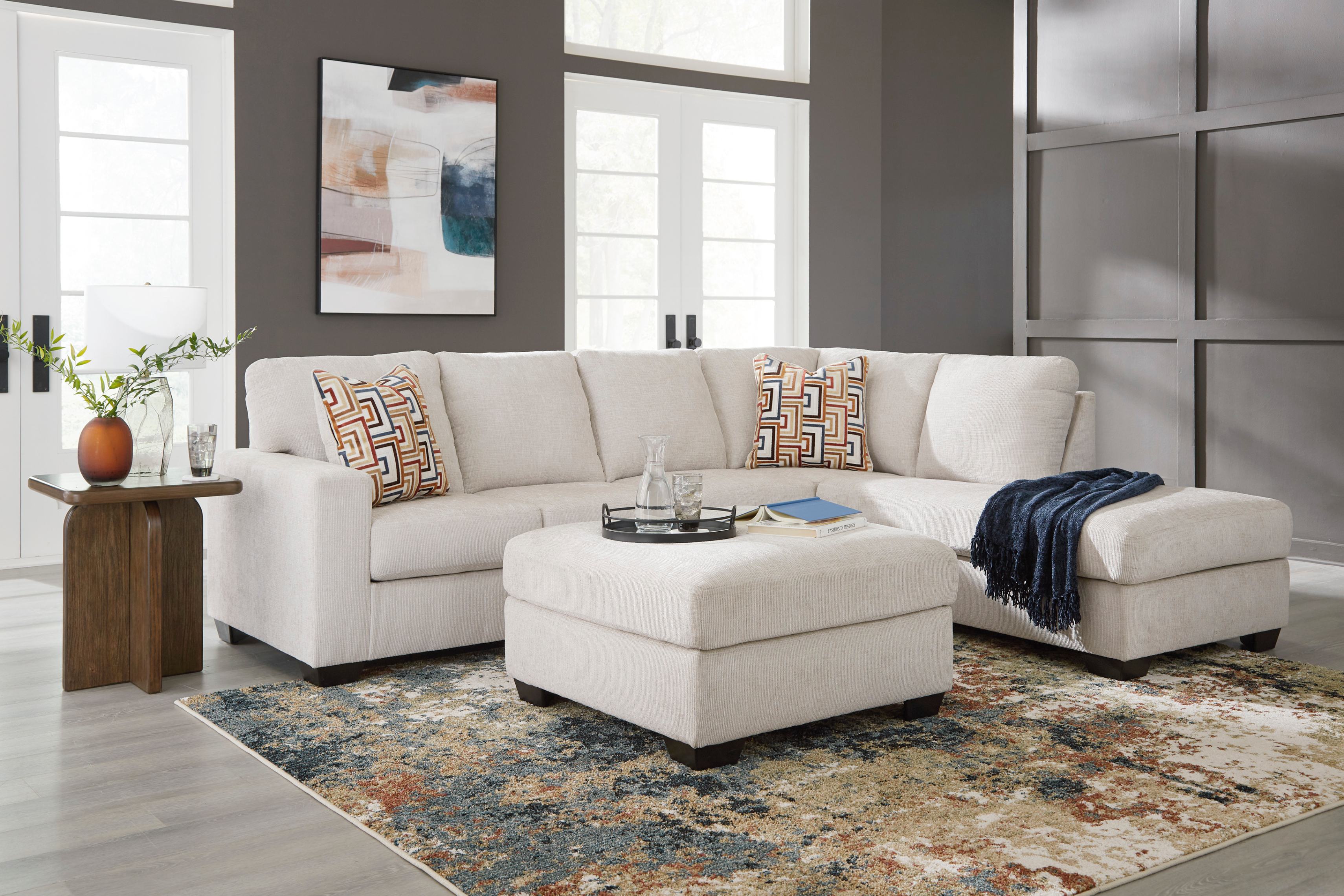 modern living room with a cream sectional sofa, patterned pillows, ottoman, blue throw, abstract art, plants, and a textured rug