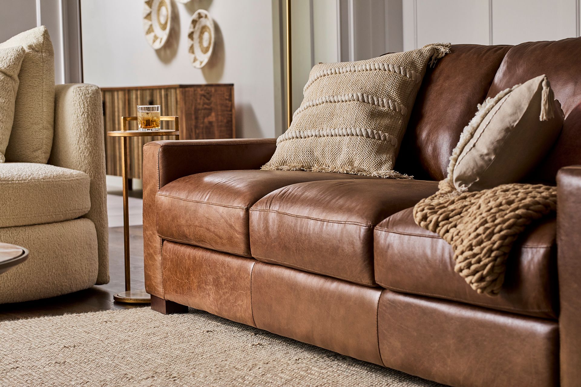 Warm living room with a brown leather sofa, textured pillows and throw, beige armchair, side table, and soft neutral décor creating a cozy tone.