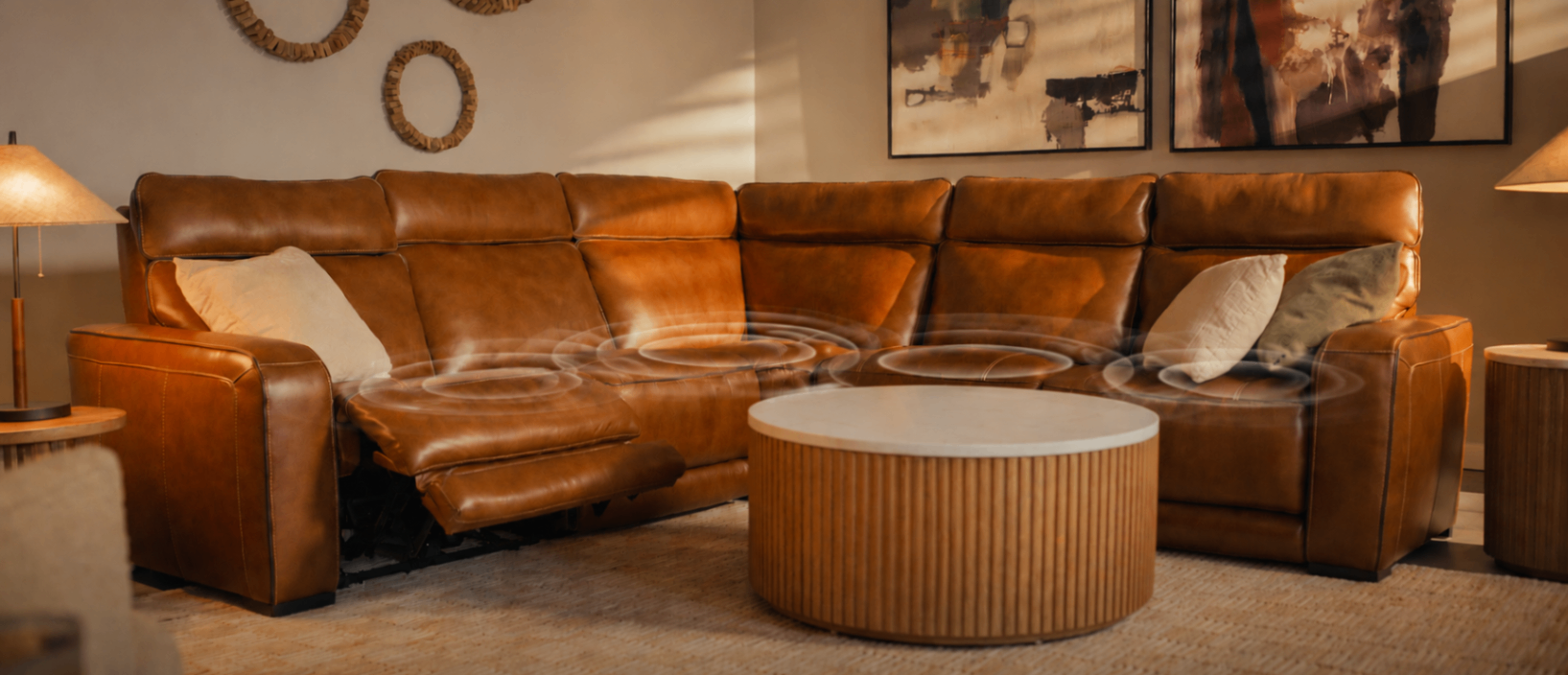 Warm living room with a large caramel leather sectional, round fluted wood coffee table, floor lamp, framed art, and cozy sunlight.