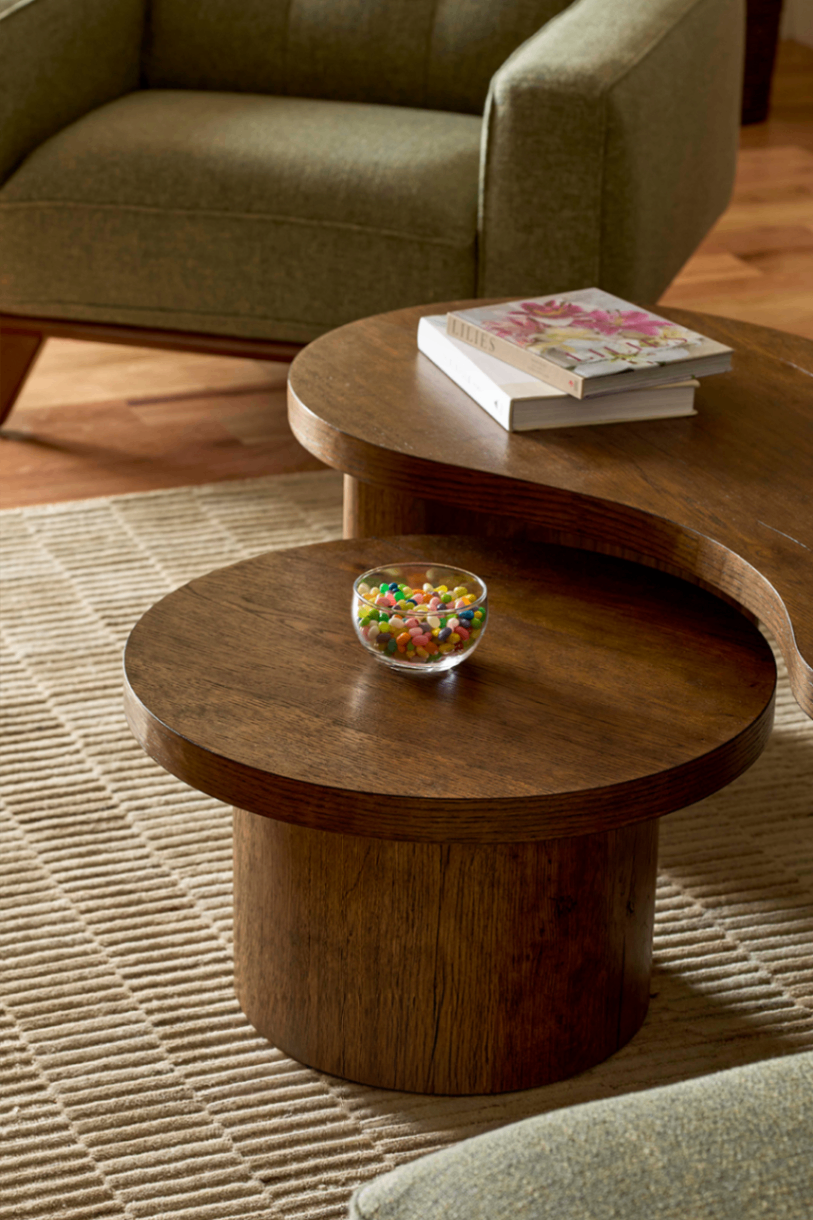 Cozy living room with nesting wooden coffee tables, a colorful candy bowl, stacked books, an armchair, and a textured rug in warm tones.