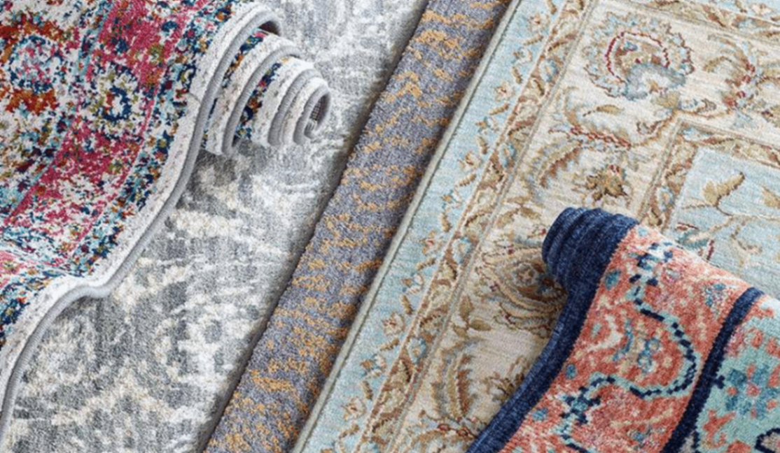 Close-up of layered patterned area rugs in cream, gray, and blue with red accents; curled edges reveal plush texture and cozy home décor.