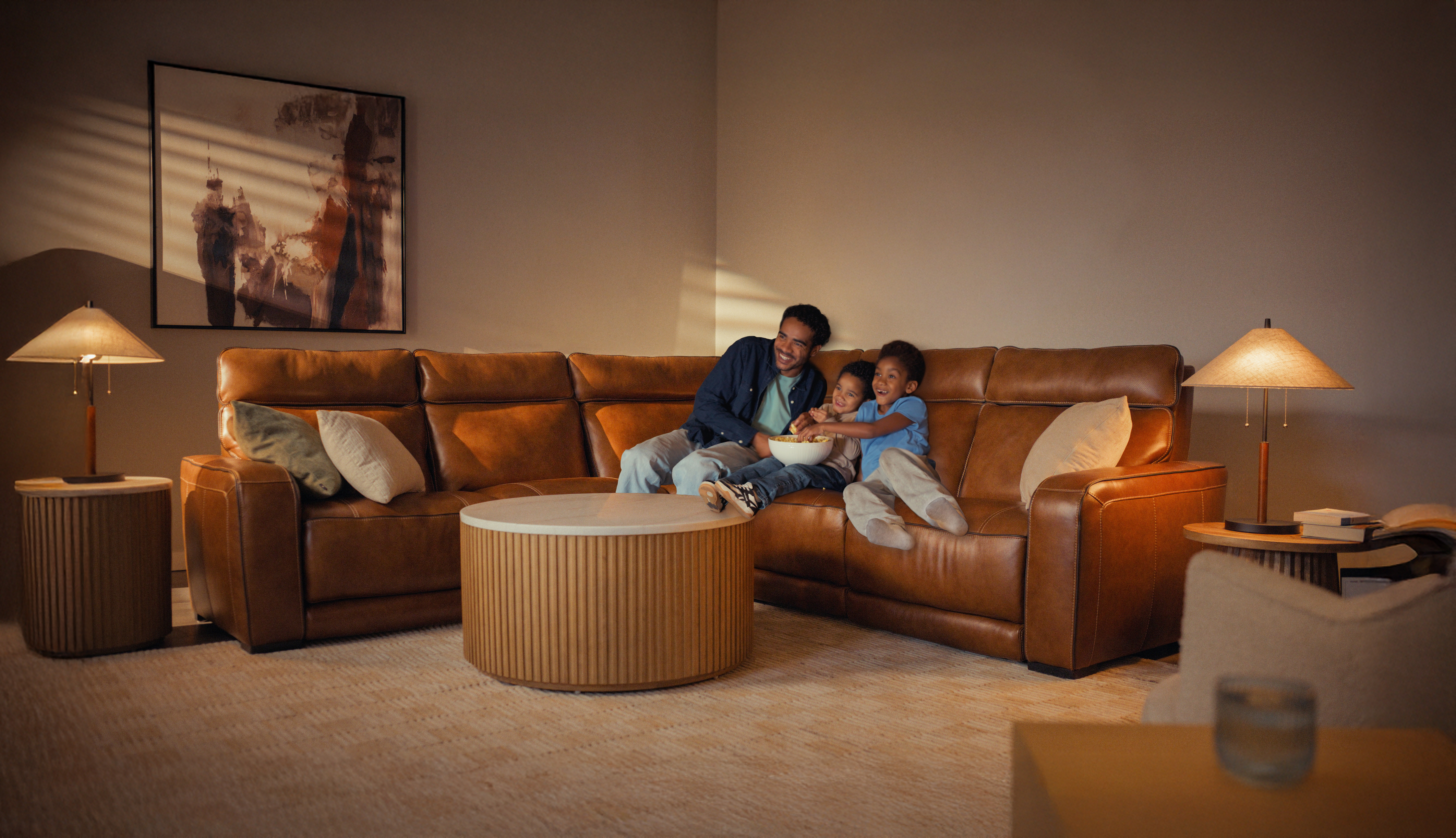 A man and two children sit on a brown leather sectional sofa, smiling and eating popcorn in a warmly lit living room.
