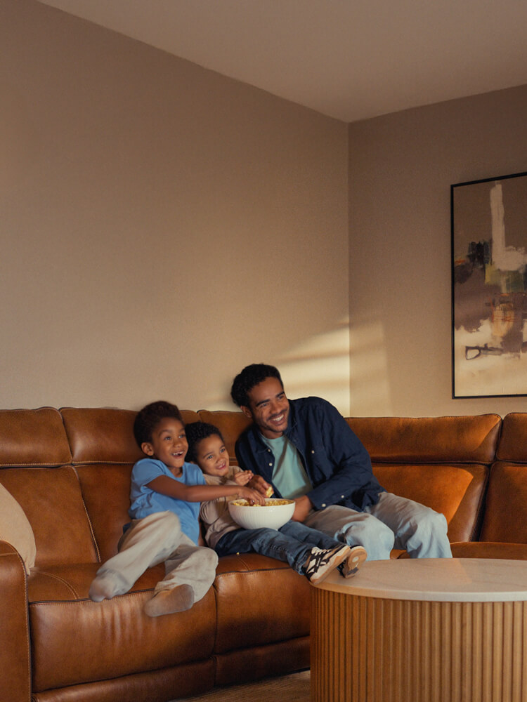 A man and two children sit on a brown leather sectional sofa, smiling and eating popcorn in a warmly lit living room.